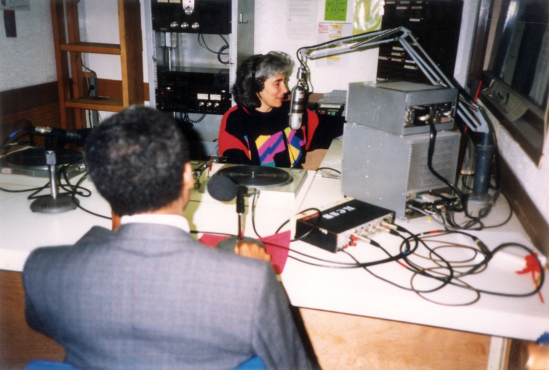 A woman wearing headphones, seated at a microphone, conducts an interview with a subject seen from behind
