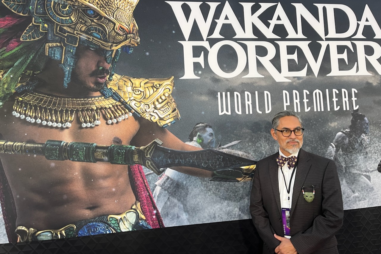 Gerardo Aldana wears a suit and stands in front of a Wakanda Forever poster which features King Namor