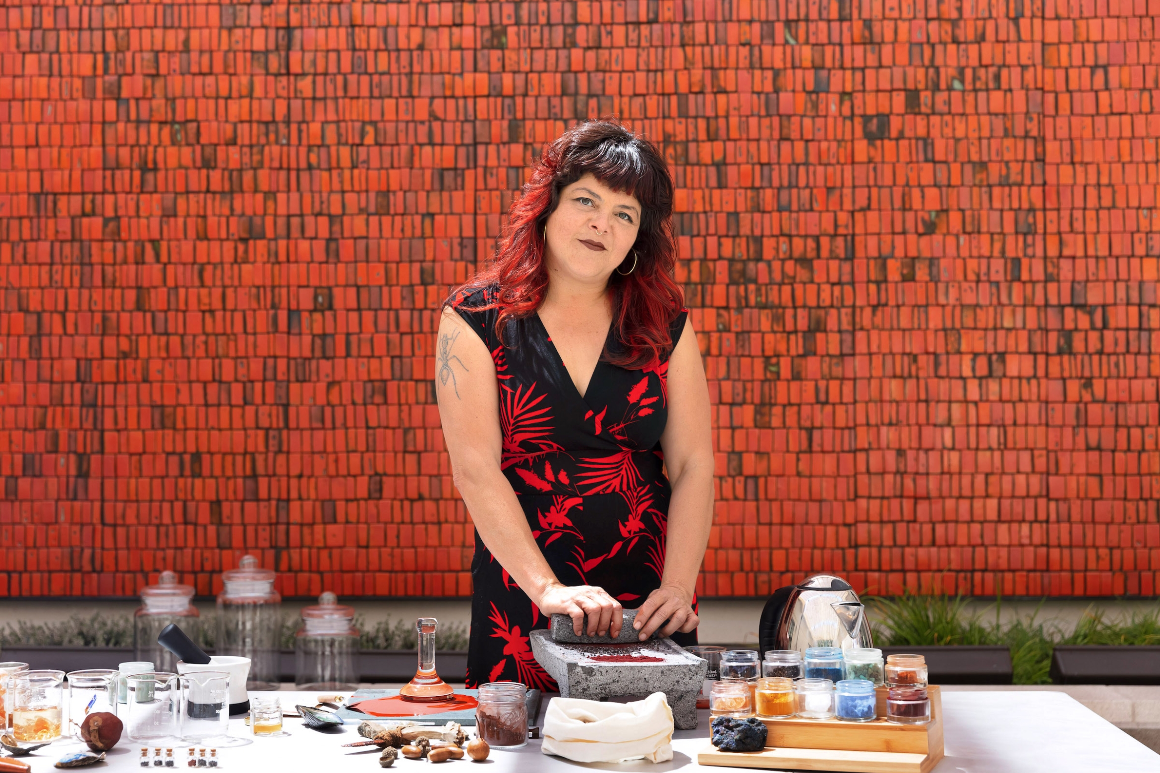 Woman with black hair with red streaks and bangs wearing a red and black dress grinds pigments on stone in front of a red tiled wall