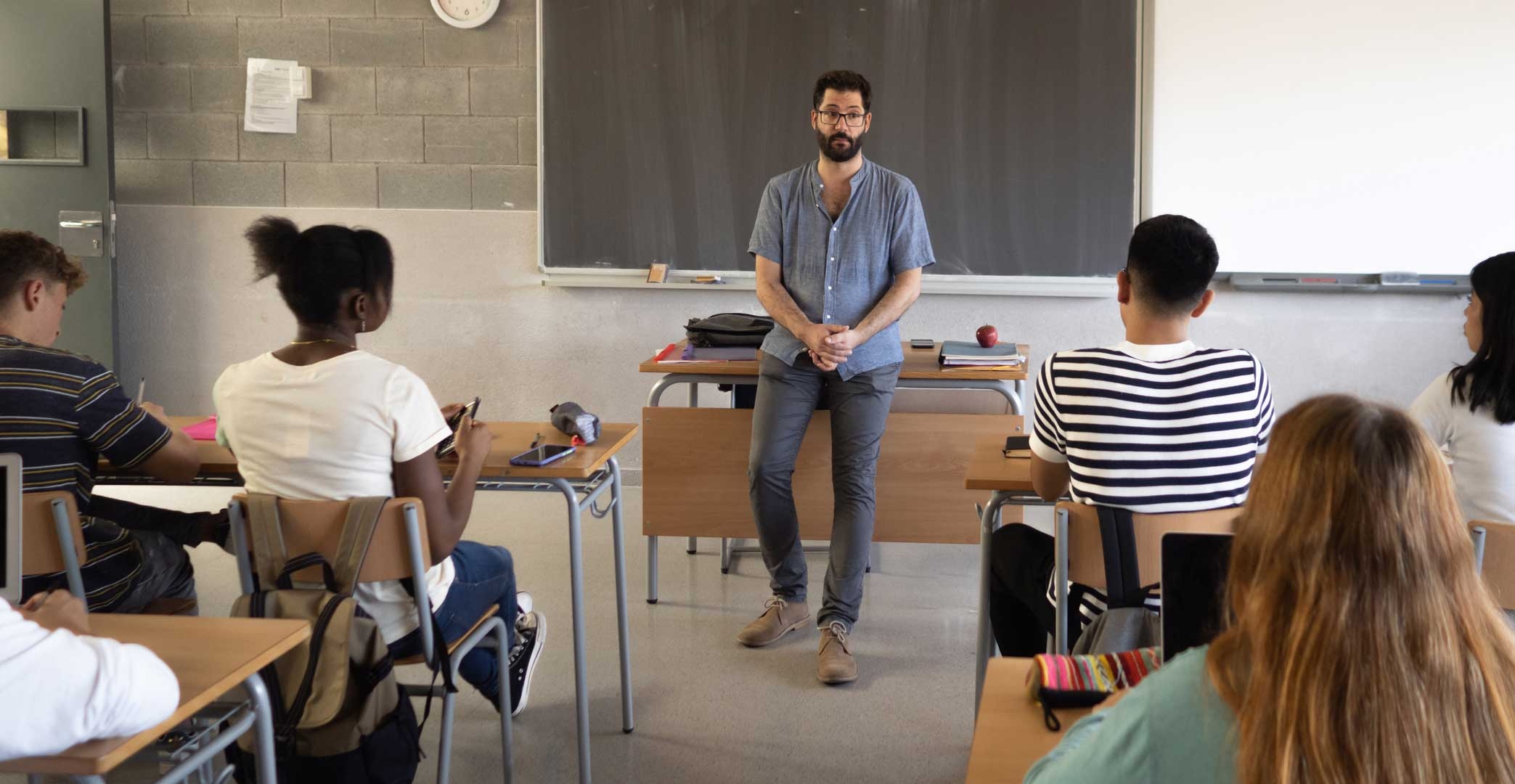 Male teacher leaning against desk talking to diverse classroom