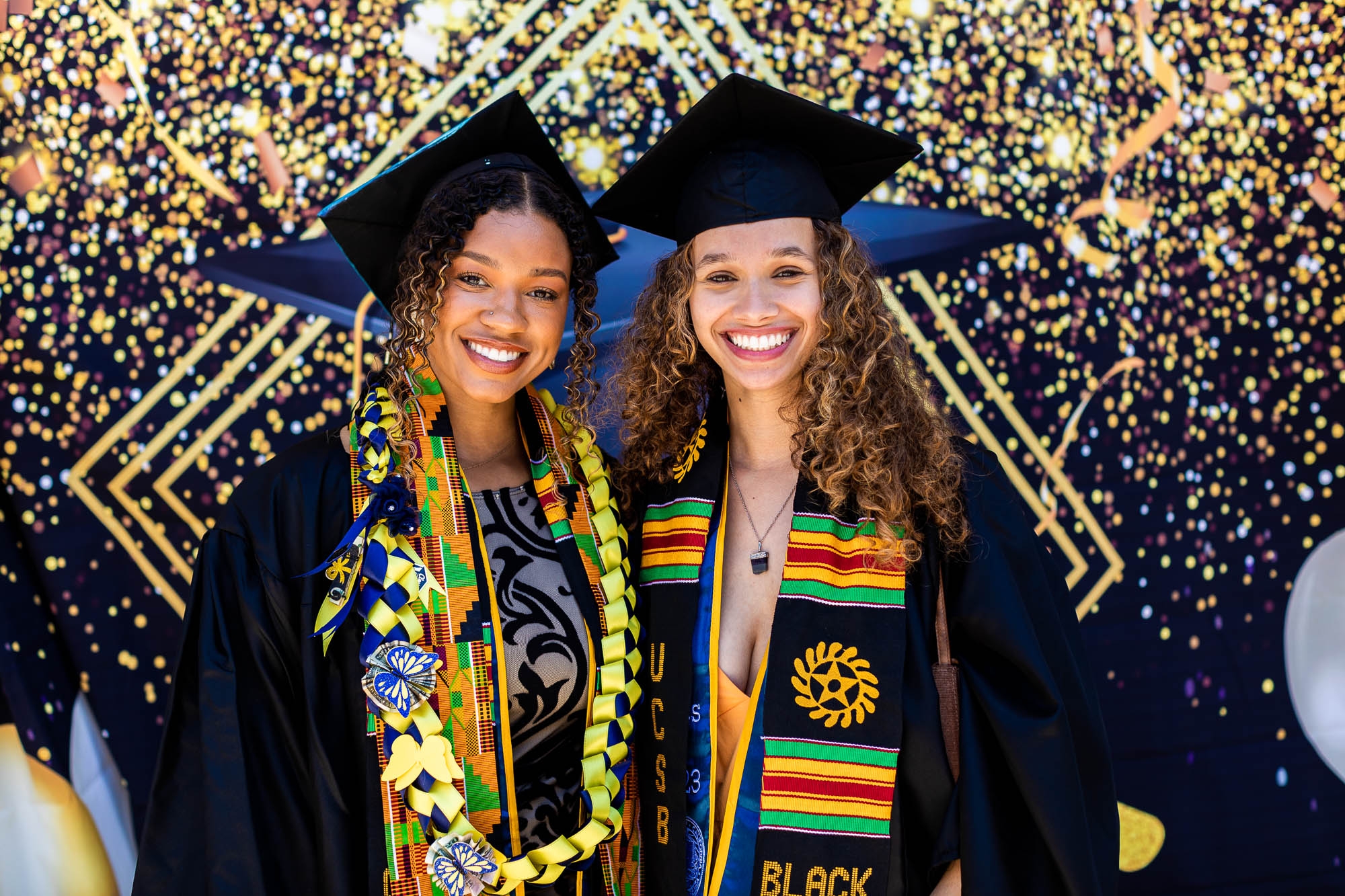 Two graduates in garb and gold glittery backdrop