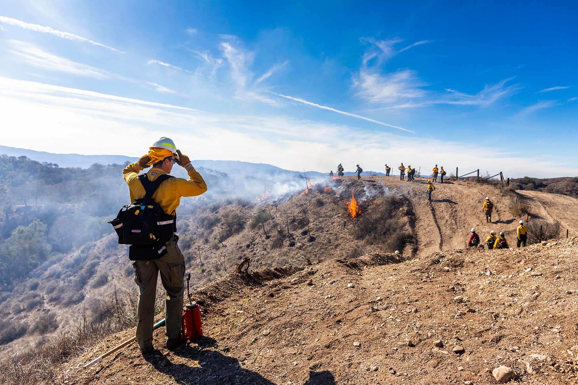 A man adjusts his hardhat on the crest of a hill as he watches the rest of the crew start the burn farther along the ridgeline.